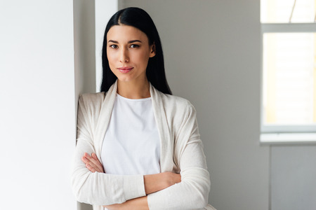 Beautiful young woman keeping arms crossed and looking at camera while standing near white wallの写真素材