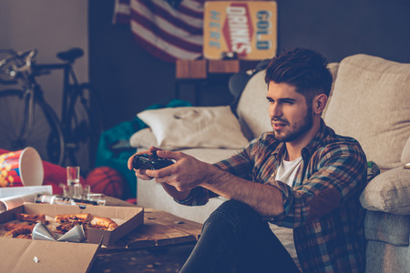 Frustrated young man holding joystick while sitting on the floor in messy room after partyの写真素材