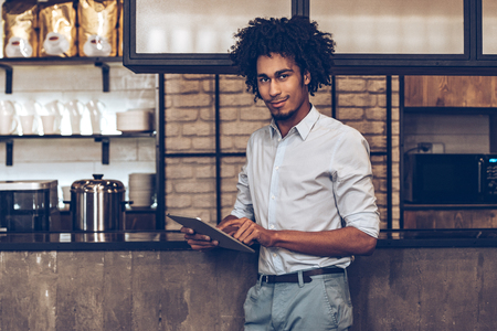 Young cheerful African man using digital tablet and looking at camera with smile while standing at bar counterの写真素材