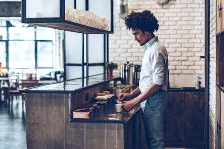 Side view of young African man stirring coffee while standing at bar counterの写真素材