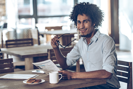 Young African man holding newspaper and looking at camera with smile while sitting in cafeの写真素材
