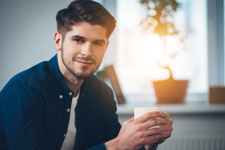 Close-up part of handsome young man holding coffee cup with smile and looking at camera while sitting on the couch at homeの写真素材