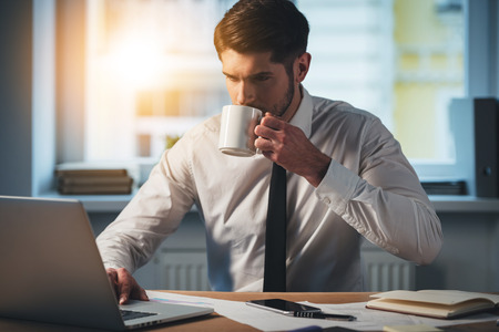 Pensive young handsome man using his laptop and drinking coffee while sitting at his working placeの写真素材