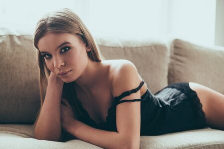 Stunning beauty. Beautiful young woman in black lingerie looking at camera while lying on the couch at homeの写真素材