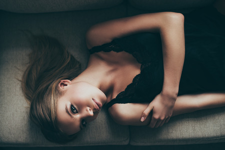 Gorgeous seductress. Top view of beautiful young woman in black lingerie looking at camera while lying on the couch at homeの写真素材