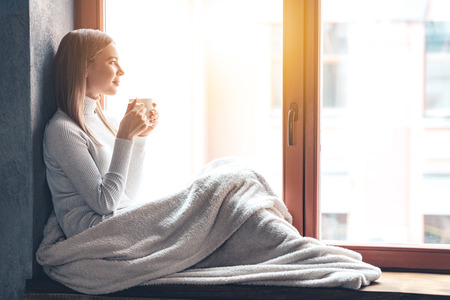 So nice to spend a day at home! Side view of beautiful young woman holding coffee cup and looking through window while sitting at windowsill at homeの写真素材