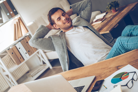 Time for little rest. Cheerful young handsome man keeping hands behind his head and eyes closed with smile while sitting at his working placeの写真素材