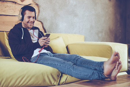 Great song! Cheerful young man in headphones using his smartphone with smile while sitting on couch at homeの写真素材