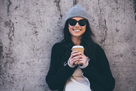 Her smile can melt your heart. Beautiful young woman in sunglasses holding coffee cup and looking at...