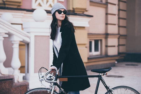 Great day for a little ride. Beautiful young woman in sunglasses rolling her bicycle and looking...