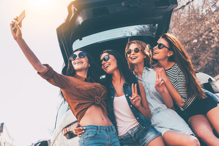 Just one selfie before road trip! Four beautiful young cheerful women making selfie with smile while standing near car trunkの写真素材