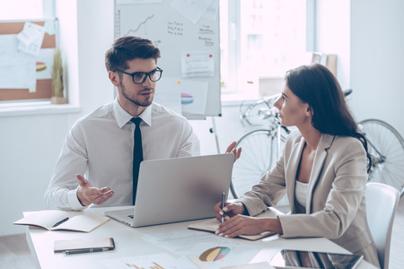 Combining their expertise. Young handsome man in glasses gesturing and discussing something with his beautiful coworker while sitting at the office tableの写真素材