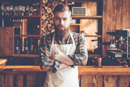 Confident in his new business. Young bearded man in apron looking at camera and keeping arms crossed while standing at bar counterの写真素材