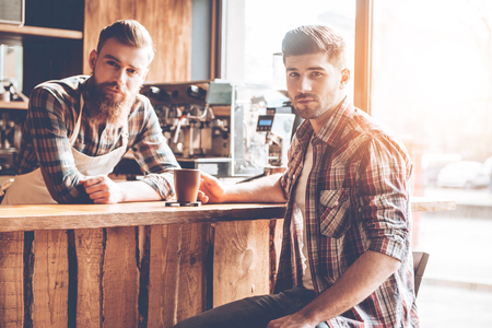Spending great time at coffee shop. Two handsome men looking at camera while sitting at bar counter at cafeの写真素材