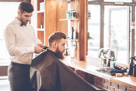 Sharp edges. Side view of young bearded man getting haircut by hairdresser with electric razor while sitting in chair at barbershopの写真素材