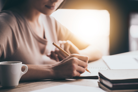 Making some notes. Close-up part of young woman writing in notebook while sitting at her working placeの写真素材