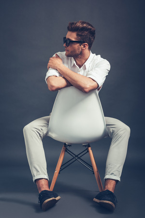 Mr Style. Full length of confident young handsome man in sunglasses looking away while sitting in chair against grey backgroundの写真素材