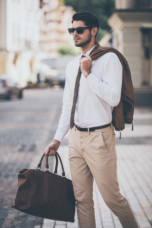 Handsome and stylish. Handsome young man in sunglasses holding leather bag and looking away while walking outdoorsの写真素材