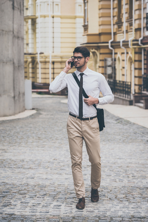 Business on a go. Confident young man in glasses talking on mobile phone while walking outdoorsの写真素材