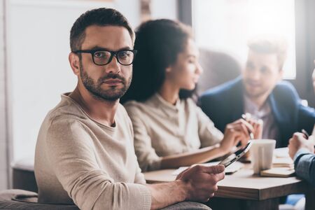 Working with his business team. Young handsome man looking at camera while sitting at the office table on business meeting with his coworkersの写真素材