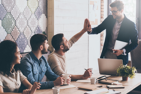We did it! Two men giving each other high-five with smile while sitting at the business meeting with their coworkersの写真素材