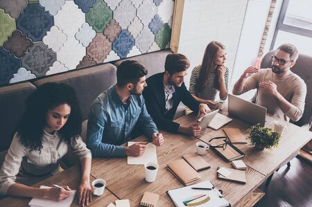 Combining their expertise. Top view of young handsome man in glasses gesturing and discussing something with his coworkers while sitting at the office tableの写真素材