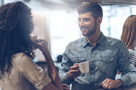 Coffee break with joy. Young handsome man holding coffee cup and discussing something with young woman while standing at bar counterの写真素材