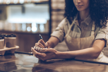 Your order. Part of young African woman in apron writing in notebook while standing at bar counterの写真素材