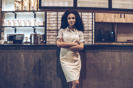 Confident cafe owner. Young cheerful African woman in apron keeping arms crossed and looking at camera with smile while standing at bar counterの写真素材