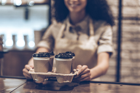 Best coffee to go! Part of young cheerful African woman in apron holding coffee cups while standing at cafeの写真素材