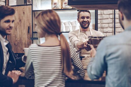 Your coffee is ready! Rear view of barista passing coffee cups to his customer while standing at bar counterの写真素材