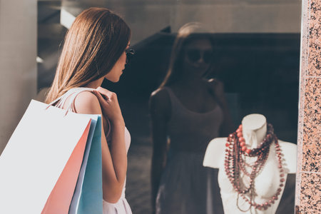 What a beautiful necklace! Rear view of beautiful young woman carrying shopping bags and looking through the window of fashion storeの写真素材