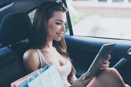 New collection in my favorite store! Beautiful young smiling woman looking at her digital tablet while sitting inside of the car with shopping bags laying near herの写真素材
