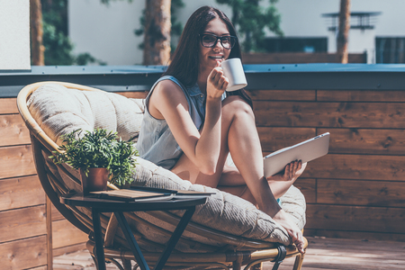 Enjoying her morning coffee. Beautiful young smiling woman holding coffee cup and looking at camera while sitting in a big comfortable chair on her outdoor house terraceの写真素材