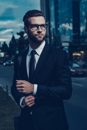 Confident young man in full suit adjusting his sleeve and looking away while standing outdoors with cityscape in the backgroundの写真素材