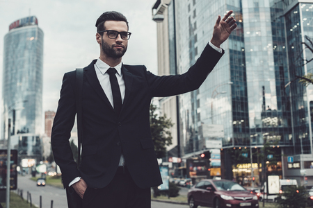 Confident young businessman in full suit catching taxi while raising his arm and standing outdoors with cityscape in the backgroundの写真素材