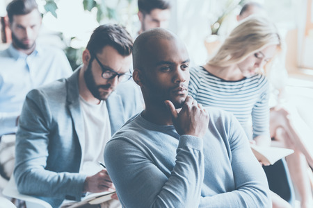 Handsome African man holding hand on chin and looking thoughtful while sitting on conference with other peopleの写真素材