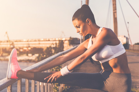 Stretching before jogging. Beautiful young woman in sports clothing doing stretching exercises while standing on the bridgeの写真素材
