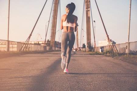 Just keep running. Full length rear view of beautiful young woman in sports clothing running along the bridgeの写真素材