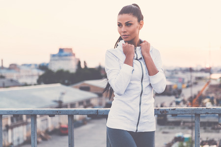 Sports style. Beautiful young woman adjusting her sports clothes and looking away while standing on the bridgeの写真素材