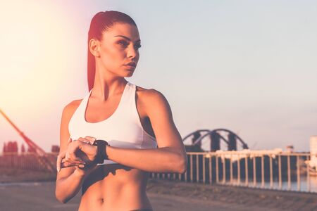 Ready to run now. Beautiful young woman in sports clothing wearing smartwatch and looking away while standing on the bridgeの写真素材