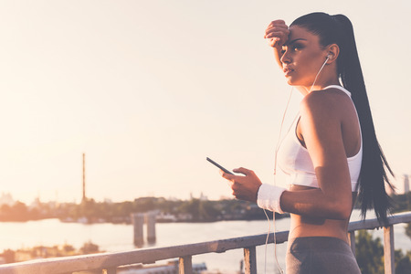Sporty woman. Beautiful young woman in headphones holding her smart phone and looking away while standing on the bridge with urban view in the backgroundの写真素材