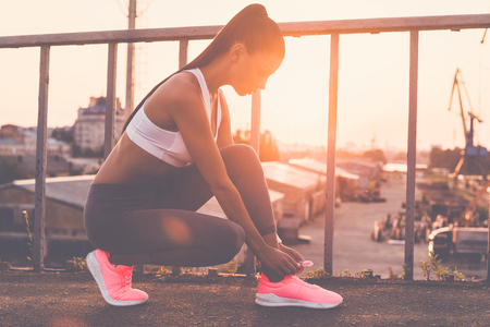 Tying her shoelaces. Beautiful young woman in sports clothing tying her shoelaces while standing on the bridge with evening sunlight and urban view in the backgroundの写真素材