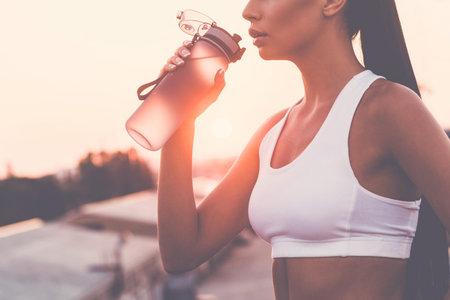 Getting refreshed. Close-up of beautiful young woman in sports clothing drinking water and looking...