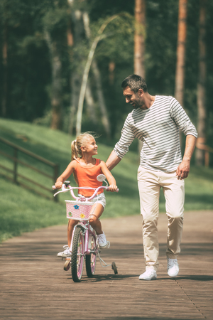 You can do it! Full length of cheerful father teaching his daughter to ride a bicycle in parkの写真素材