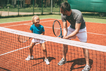 Tennis training. Cheerful father in sports clothing teaching his daughter to play tennis while both standing on tennis courtの写真素材