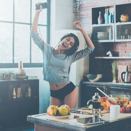 Kitchen dance. Beautiful young mixed race woman in headphones dancing while standing in kitchen at homeの写真素材