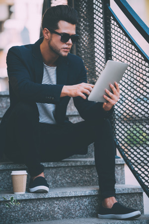 Surfing web outdoors. Handsome young man in smart casual wear working on digital tablet while sitting on stairs outdoorsの写真素材