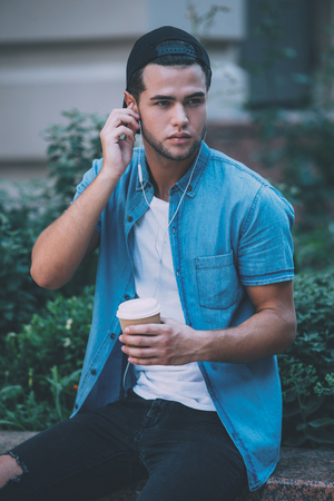 Enjoying favorite music and fresh coffee. Handsome young man adjusting his headphones and looking away while holding coffee cup and sitting outdoorsの写真素材