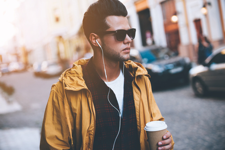 Enjoying city life. Handsome young man in headphones carrying coffee cup while walking along the city streetの写真素材